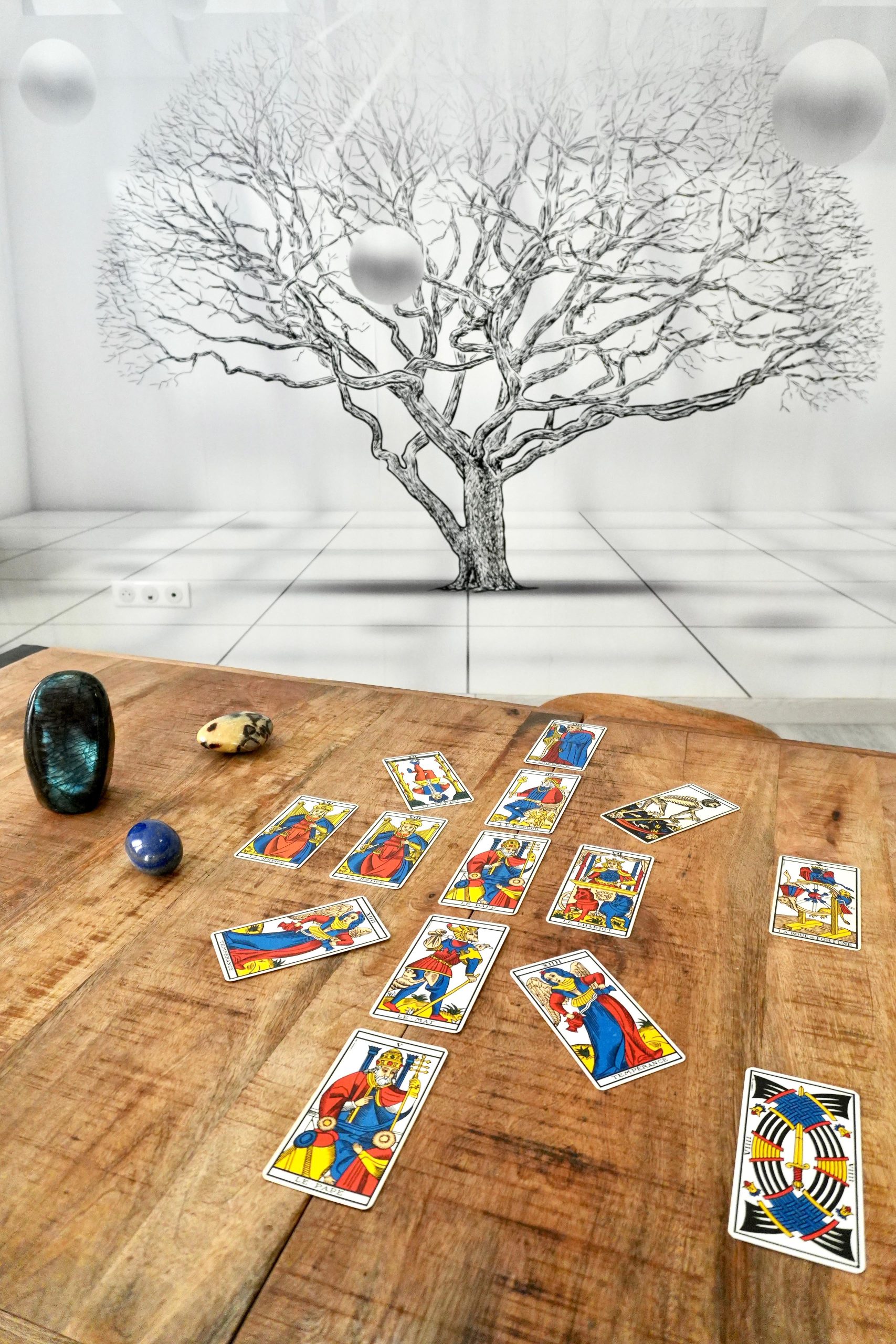 Référentiel de Naissance posé sur une table en bois, composé de cartes du Tarot de Marseille disposées selon une figure symbolique. En arrière-plan, un grand arbre noir et blanc représentant l’arbre généalogique évoque les liens transgénérationnels, accompagné de pierres minérales dans un espace de consultation lumineux.